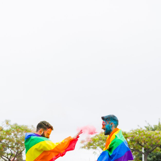 Hombres alegres en banderas de arco iris tirando pintura en polvo en la calle