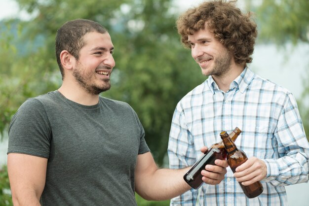 Hombres alegres que tintinean botellas de cerveza en la naturaleza