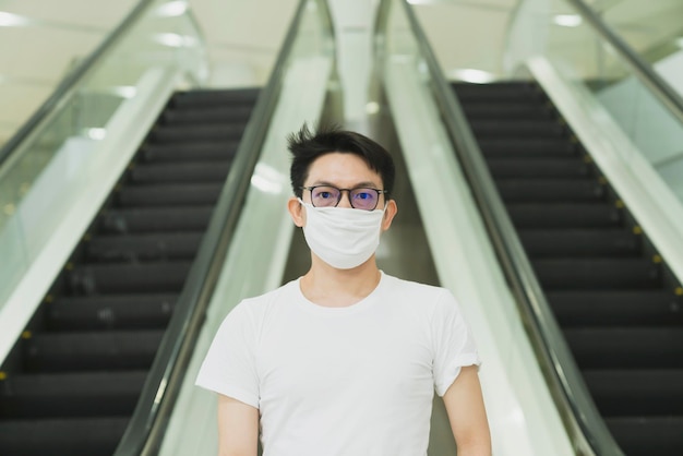 Los hombres asiáticos jóvenes usan una máscara blanca protectora para evitar el virus de la corona o el covid 19 propagan el hombre asiático de pie junto al estilo de vida urbano de la escalera mecánica