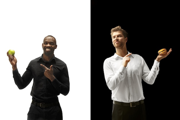 Hombres comiendo una hamburguesa y frutas frescas sobre un fondo blanco y negro. Los felices hombres afro y caucásicos. La hamburguesa, el concepto de comida rápida, saludable y no saludable.