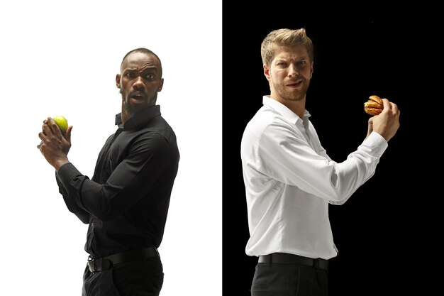 Hombres comiendo una hamburguesa y frutas frescas sobre un fondo blanco y negro. Los felices hombres afro y caucásicos. La hamburguesa, el concepto de comida rápida, saludable y no saludable.