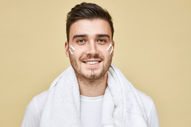 Hombres, concepto de estilo de vida, belleza y cuidado de la piel. Imagen de joven feliz con cerdas posando aislado con rayas de espuma en las mejillas y toalla de baño alrededor del cuello, va a lavarse la cara y afeitarse la barba