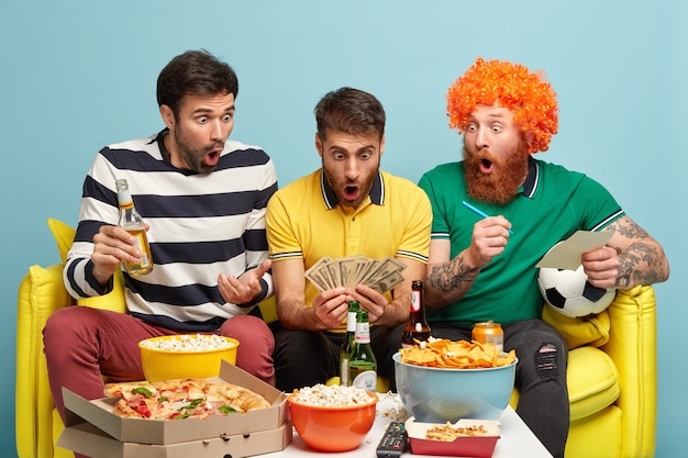 Foto gratuita los hombres estupefactos miran sorprendentemente el dinero en efectivo, emocionados de ganar una apuesta, ven partidos de fútbol en la televisión, participan en juegos de azar, comen comida rápida. chico emocional con mucho dinero, disfruta de la liga de campeones.