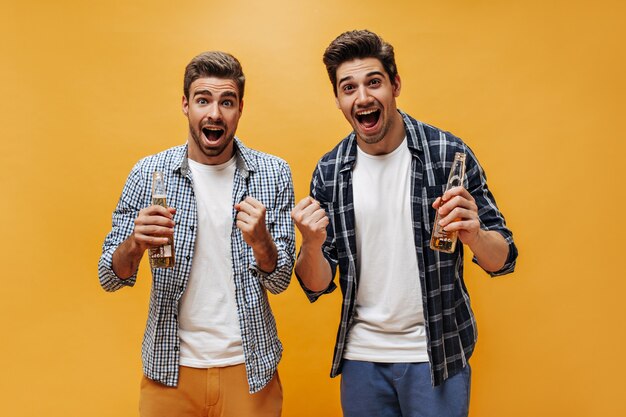 Hombres felices emocionados sorprendidos con camisetas blancas y camisas a cuadros se regocijan, miran a la cámara y sostienen botellas de cerveza en la pared naranja.