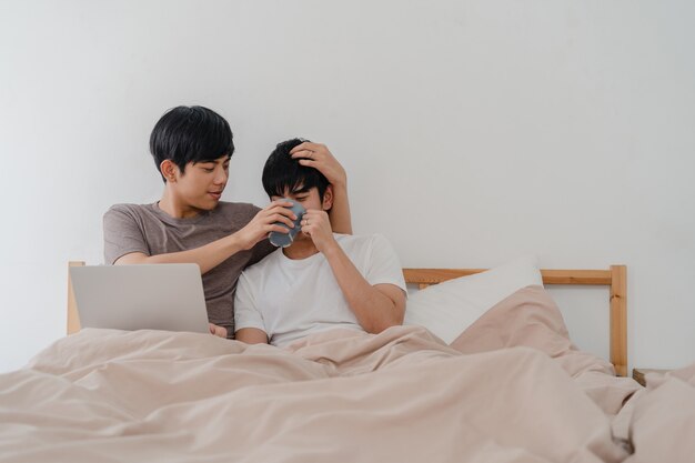 Los hombres gay asiáticos se juntan usando la computadora portátil de la computadora y bebiendo café en el hogar moderno Joven amante de Asia masculino feliz relajarse descansar juntos después de despertarse, viendo la película acostada en la cama en el dormitorio en la casa en la mañana