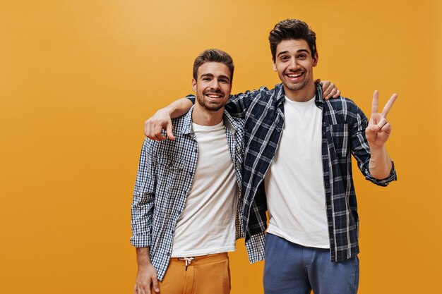 Hombres jóvenes alegres con camisas azules a cuadros, camisetas blancas y pantalones coloridos posan en la pared naranja de buen humor y sonríen.