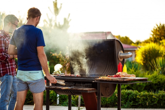 Hombres jovenes que asan la barbacoa en parrilla en campo de la cabaña.