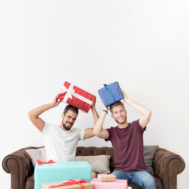 Hombres jovenes sonrientes que se sientan en el sofá que sostiene las cajas de regalo en su cabeza