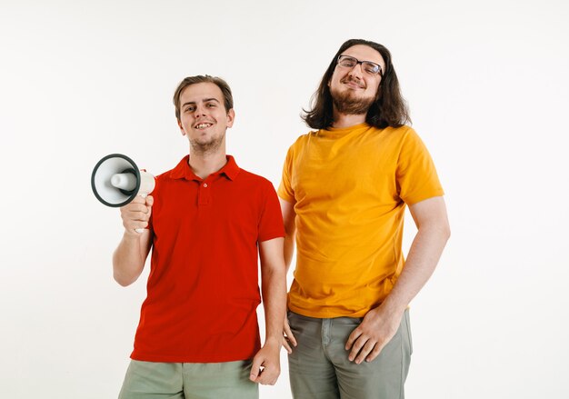 Los hombres jóvenes vestían los colores de la bandera LGBT en una pared blanca. Modelos masculinos caucásicos en camisas brillantes. Luce feliz, sonriente y abrazado. Orgullo LGBT, derechos humanos y concepto de elección. Sosteniendo la boquilla.