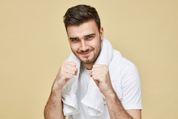 Hombres y masculinidad. Retrato de hombre sin afeitar morena joven confiado con una toalla alrededor del cuello de pie en el baño sosteniendo los puños cerrados frente a él, desafiando su propio reflejo en el espejo