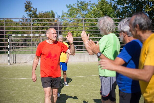 Hombres mayores activos agradeciéndose unos a otros después del partido. Hombres con cabello gris en ropa deportiva parados en fila en el campo deportivo, dando cinco años. Fútbol, deporte, concepto de ocio.