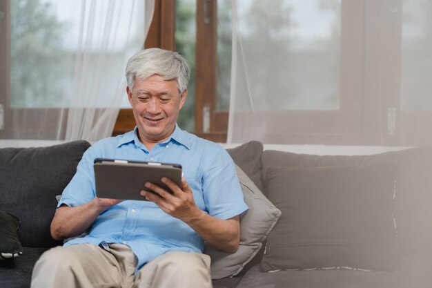 Hombres mayores asiáticos que usan la tableta en casa. Información de búsqueda masculino chino mayor asiático sobre cómo buena salud en internet mientras se está acostado en el sofá en concepto de sala de estar en casa.