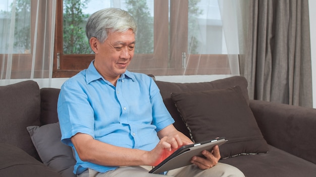 Hombres mayores asiáticos que usan la tableta en casa. Información de búsqueda masculino chino mayor asiático sobre cómo buena salud en internet mientras se está acostado en el sofá en concepto de sala de estar en casa.
