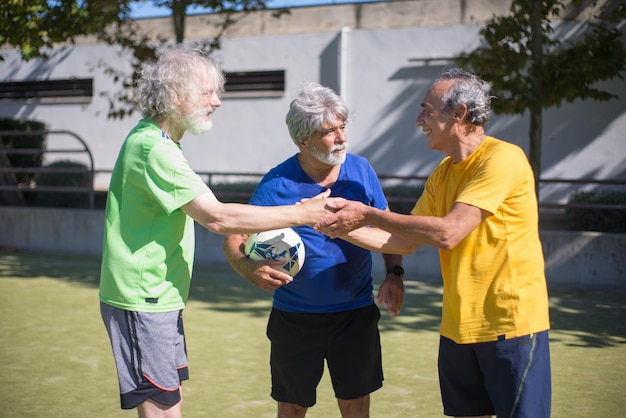 Hombres mayores entusiastas agradeciéndose unos a otros después del partido. Hombres con cabello gris en ropa deportiva de pie en el campo deportivo, dándose la mano. Fútbol, deporte, concepto de ocio.