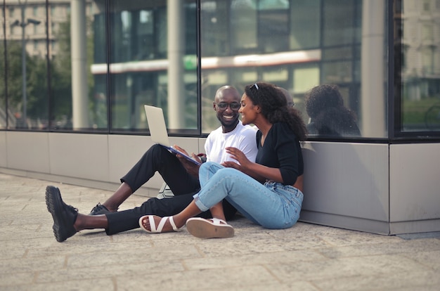 Hombres y mujeres africanos sonrientes alegres sentados en el suelo y usando una computadora portátil durante el día