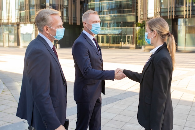 Hombres y mujeres de negocios en mascarillas y trajes de oficina reunidos en la ciudad, dándose la mano cerca del edificio. Tiro de vista lateral. Concepto de comunicación y protección antivirus