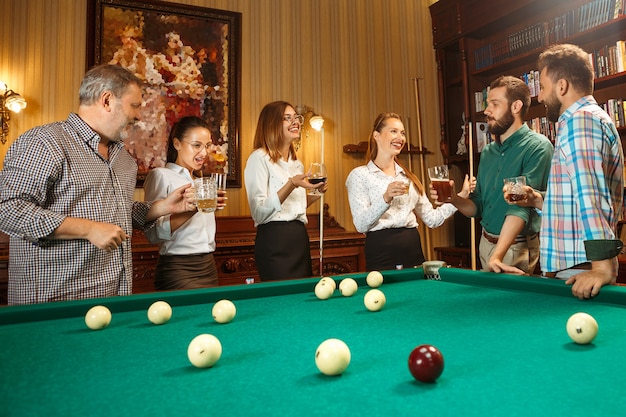 Hombres y mujeres sonrientes jovenes que juegan al billar en la oficina o en casa después del trabajo. Compañeros de trabajo involucrados en actividades recreativas. Amistad, actividad de ocio, concepto de juego.
