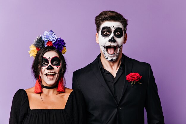 Hombres y mujeres traviesos alegres en ropa negra con detalles en rojo gritan de asombro, posando con maquillaje de Halloween para el retrato.