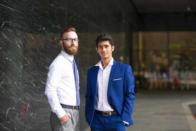 Foto gratuita hombres multiétnicos jovenes serios que se colocan cerca del edificio de oficinas