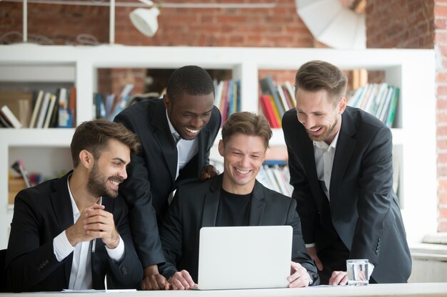 Hombres de negocios sonrientes multiétnicos en trajes que miran algo divertido en el ordenador portátil