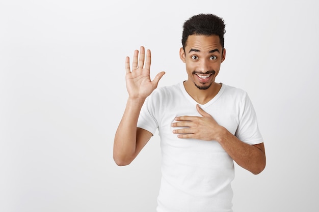 Honesto y sincero chico guapo de piel oscura que hace una promesa, sostiene la mano en el corazón y un brazo levantado, jura decir la verdad