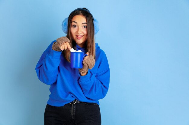 Hora de cafe. Retrato de mujer caucásica sobre fondo azul de estudio. Modelo de mujer hermosa en ropa de abrigo. Concepto de emociones, expresión facial, ventas, publicidad. Estado de ánimo de invierno, Navidad, vacaciones.