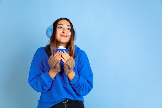 Hora de cafe. Retrato de mujer caucásica sobre fondo azul de estudio. Modelo de mujer hermosa en ropa de abrigo. Concepto de emociones, expresión facial, ventas, publicidad. Estado de ánimo de invierno, Navidad, vacaciones.