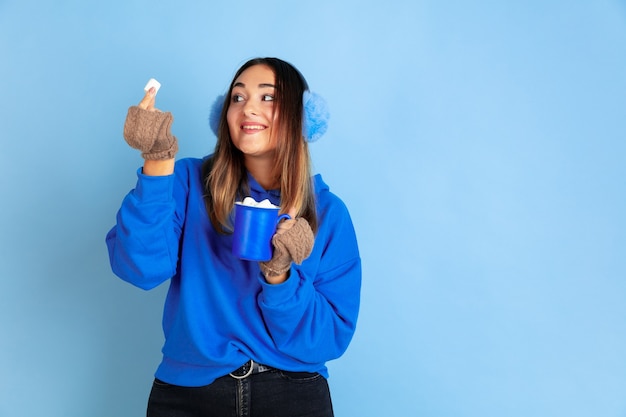 Hora de cafe. Retrato de mujer caucásica sobre fondo azul de estudio. Modelo de mujer hermosa en ropa de abrigo. Concepto de emociones, expresión facial, ventas, publicidad. Estado de ánimo de invierno, Navidad, vacaciones.