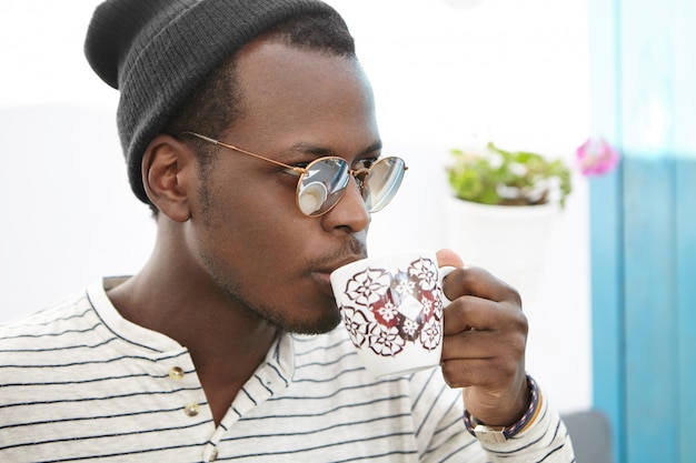 Hora de cafe. Seguros de moda hombre africano con sombrero y sombras sosteniendo la taza, bebiendo capuchino fresco, mirando hacia delante con expresión pensativa, disfrutando de una bebida caliente durante el almuerzo en el café