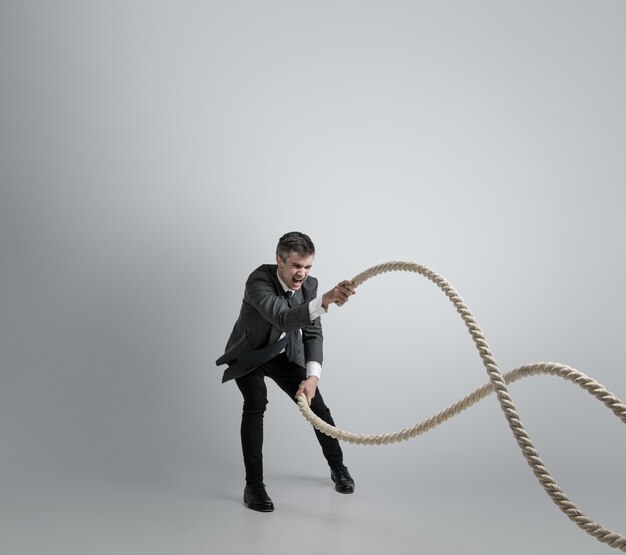 Hora de la fuerza. Hombre en ropa de oficina entrenando con cuerdas en la pared gris. Consigue objetivo, superando problemas, plazos. Hombre de negocios en movimiento, acción. Deporte, estilo de vida saludable, trabajo.