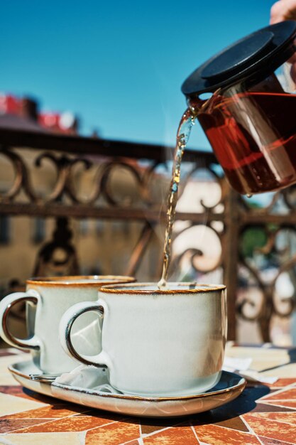 La hora del té por la mañana en la antigua veranda El té rojo en una tetera de vidrio se vierte en tazas de tiro vertical Vacaciones de verano ambiente de fiesta de té descanso y relajación Enfoque selectivo en una taza