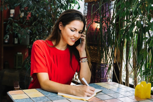 Horario hermoso de la escritura de la mujer en diario mientras que habla en el teléfono móvil