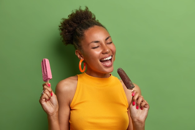 Horario de verano, ocio, diversión, concepto de alimentación. Feliz adolescente étnica hace el tonto, canta en voz alta y come helado de chocolate stawberry. Mujer con bocadillo congelado dulce delicioso fresco azucarado