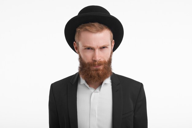 Horizontal guapo elegante joven pelirrojo masculino con barba difusa, frunciendo los ojos y frunciendo los labios, con mirada sospechosa. Hombre sin afeitar con sombrero y traje disgustado y enojado