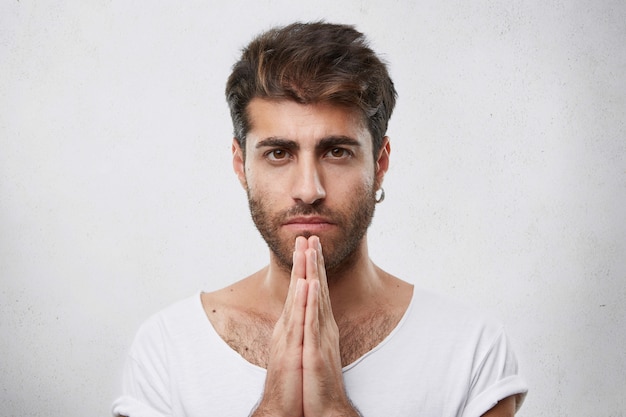 Foto gratuita horizontal retrato de hombre guapo con peinado de moda y barba con aretes y camiseta blanca manteniendo las manos juntas rezando con ojos llenos de creer para pedir algo mejor