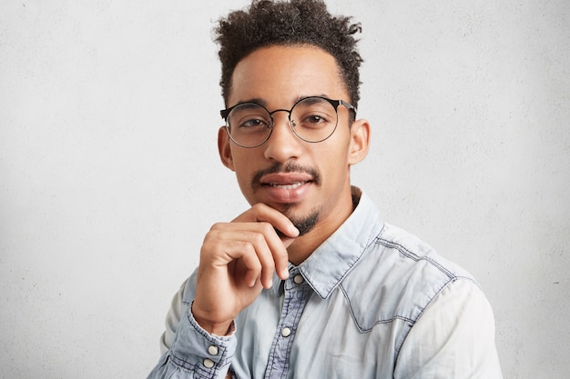 Horizontal retrato de hombre de piel oscura con peinado moderno, viste camisa de mezclilla y gafas redondas