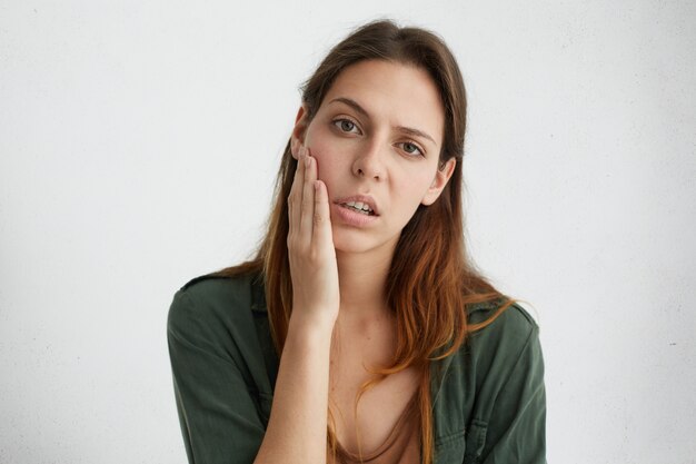 Horizontal retrato de mujer bastante triste con brillantes ojos oscuros y cabello lacio sosteniendo la mano en la mejilla mirando molesto