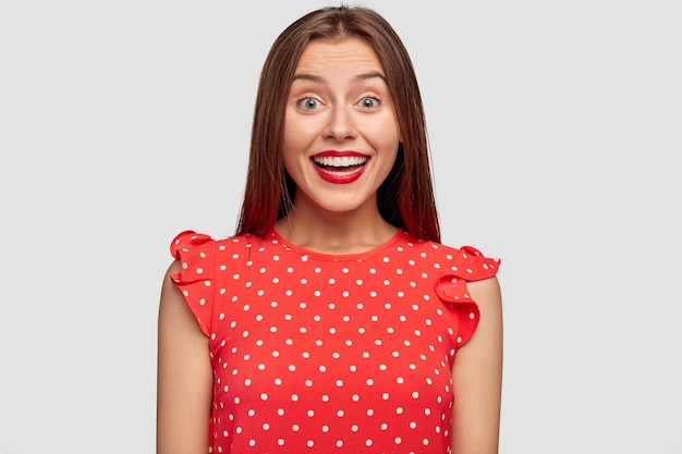 Horizontal retrato de mujer encantadora con lápiz labial rojo posando contra la pared blanca