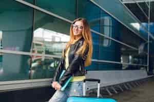Foto gratuita horizontal retrato de niña bonita con pelo largo parado afuera en el aeropuerto. viste chaqueta negra con jeans, sostiene una computadora portátil en la mano. ella sonríe a la cámara.