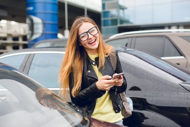 Horizontal retrato de niña con cabello largo con gafas caminando en la zona de estacionamiento. Viste suéter amarillo y chaqueta negra. Ella está sonriendo a la cámara y sostiene el teléfono en las manos.