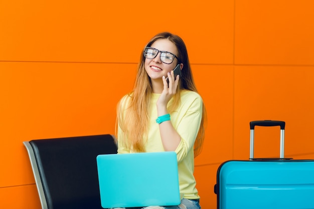 Horizontal retrato de niña en suéter amarillo sentado en una silla sobre fondo naranja. Tiene el pelo largo y gafas negras. Ella está hablando por teléfono y sonriendo a la cámara.