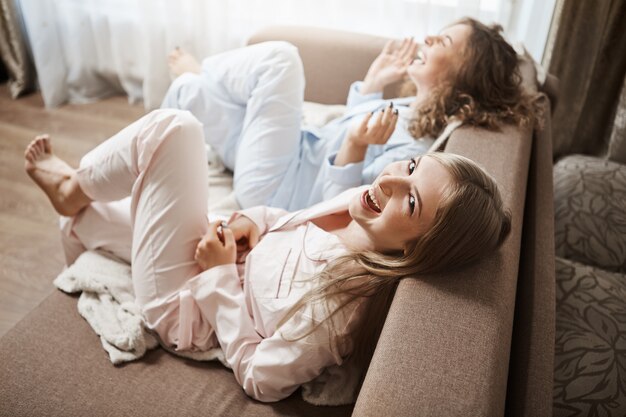 Los huesos perezosos no hacen nada y disfrutan del ocio juntos. Tiro de ángulo superior de lindas mujeres sentadas en el sofá en ropa de dormir riendo a carcajadas, hablando de momentos incómodos en la oficina, pasando los fines de semana