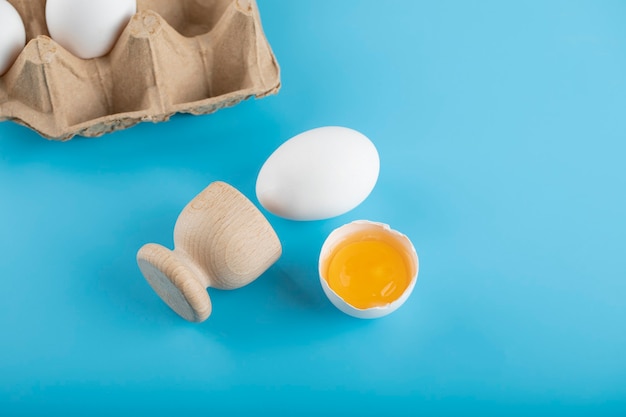 Huevo crudo roto y contenedor de huevos en superficie azul.