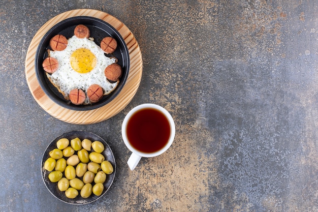 Huevo frito y salchichas en una sartén servido con aceitunas y té