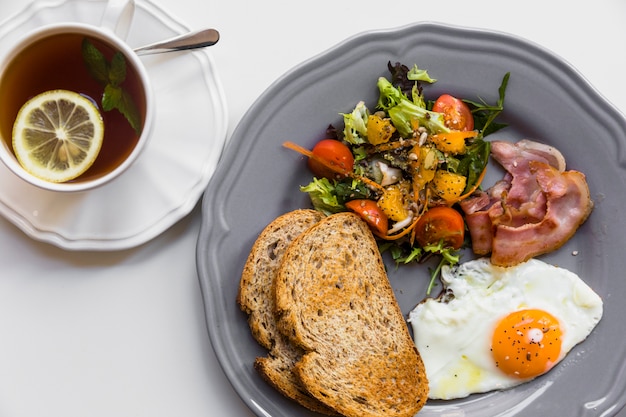 Huevo medio frito; brindis; ensalada; tocino en placa gris con limón y menta taza de té sobre fondo blanco