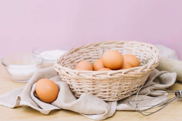 Huevos en canasta de mimbre y batidores en mesa de madera.