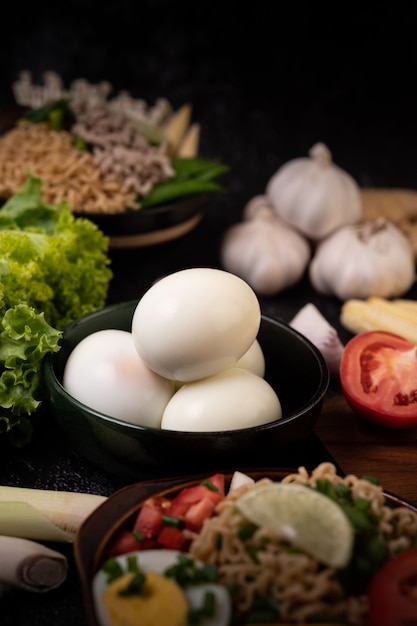 Huevos duros en un tazón negro, ajo, salchicha, tomates colocados sobre una tabla de cortar de madera