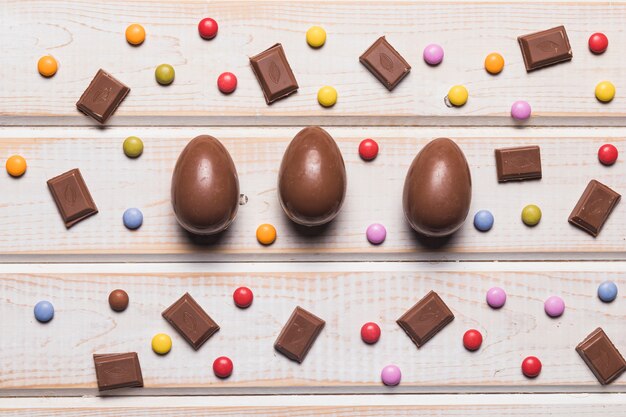 Huevos enteros de chocolate de pascua; Pedazos y gemas de colores caramelos multicolores en escritorio de madera