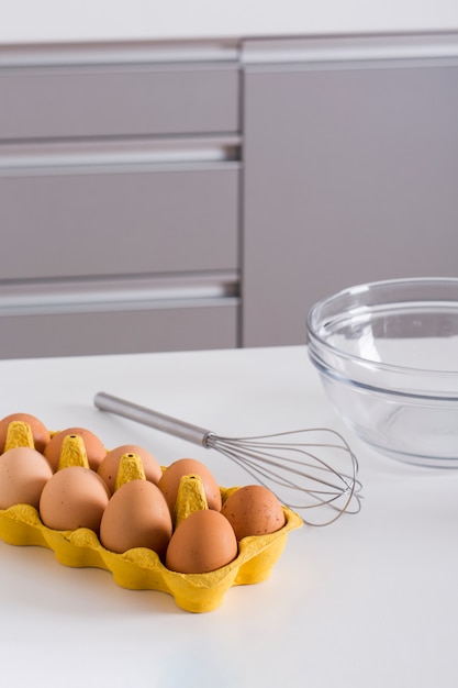 Foto gratuita huevos de la frente en el cartón amarillo; batidor y recipiente de vidrio en mesa blanca