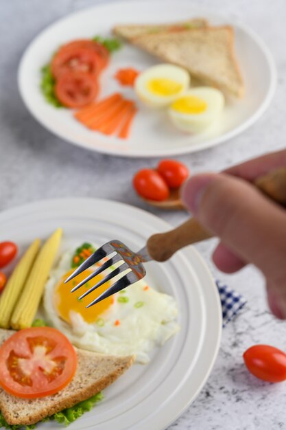 Huevos fritos, pan, zanahorias y tomates en un plato blanco para el desayuno, enfoque selectivo de mano con un tenedor.